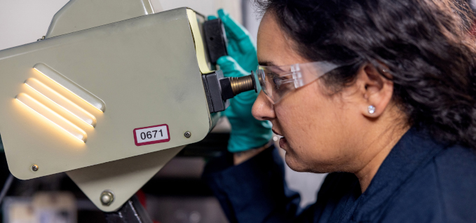 A side portrait of a lab technician wearing safety glasses and protective gloves looking through a microscope.