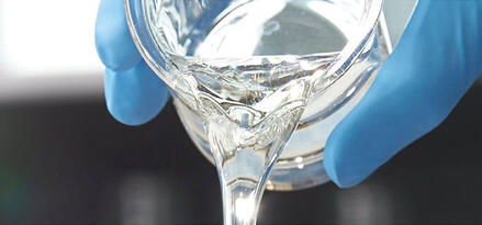 A close-up shows a clear, viscous liquid being poured from a glass beaker. The beaker is held by gloved hands wearing bright blue nitrile gloves, suggesting a lab or cleanroom setting. The liquid forms a smooth stream with glossy reflections. The background is softly blurred, keeping focus on the pouring action and laboratory glassware.