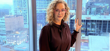 A person with curly blonde hair is standing indoors near large windows overlooking a cityscape. The background features several modern high-rise buildings with glass facades and some older, brick structures. The person is wearing a dark blouse with a bow collar and has one hand resting on the window frame. The lighting appears natural, with daylight streaming in, giving the room a cool, urban atmosphere. The overall setting suggests a metropolitan environment, possibly in an office or apartment.