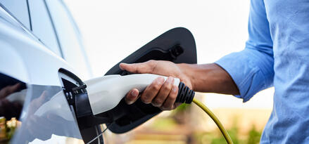 The image shows a close-up of a person’s hand plugging a charging cable into an electric vehicle. The individual is wearing a light blue long-sleeve shirt, and the cable is yellow with a white and black charging connector. The vehicle’s charging port is open and clearly visible, with the connector being inserted. The background is slightly blurred, featuring greenery and a fence, indicating the scene is likely outdoors, possibly at a home or a charging station. The focus is on the act of charging the electric vehicle, highlighting sustainable transportation.