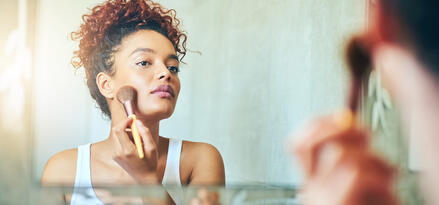A person is standing in front of a bathroom mirror, applying makeup with a brush. They are wearing a white tank top and have curly hair pulled up. The background shows a light-colored wall and part of a shower fixture, indicating the setting is a bathroom. The scene is softly lit, with sunlight streaming in from the left side, creating a warm atmosphere.