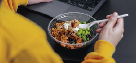 The image shows a person wearing a bright yellow sweater working on a laptop while eating from a clear plastic bowl. The bowl contains a colorful meal, including edamame beans, some type of grain or rice, pieces of what looks like sweet potatoes or another orange vegetable, nuts, and a dollop of white sauce. The person is using a white plastic fork to eat. The scene suggests a casual work or study environment, possibly during a lunch break. The focus is on multitasking—working and eating at the same time.