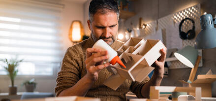 A person is working on assembling a small architectural model, holding a bottle of glue and applying it to the model. The workspace is well-lit with natural light coming from a window and additional desk lamps. The table is scattered with other model components, indicating ongoing creative work. The background includes shelves, potted plants, headphones, and pegboard with tools, suggesting a home studio or workshop environment. The overall atmosphere is cozy and organized, suitable for crafting or design activities.