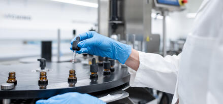 The image shows a person wearing a white lab coat and blue nitrile gloves working in a laboratory setting. They are using a dropper to add liquid to small amber glass bottles, which are positioned on a rotating tray or carousel, likely part of a laboratory automation or filling system. The background features clean, modern laboratory equipment and a digital screen, indicating a controlled and sterile environment typical of pharmaceutical or scientific research labs. The focus is on precision and careful handling of the substances being dispensed.
