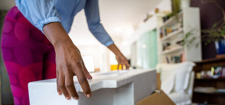 The image shows a person unpacking a cardboard box in a home setting. The individual is lifting a piece of white foam packaging, suggesting that a fragile or valuable item was inside the box. The person is wearing a light blue shirt and vibrant magenta pants with a circular pattern. In the background, a living room is visible, featuring a bookshelf, indoor plants, and a white chair, creating a cozy atmosphere. The scene conveys a moment of settling in, possibly after moving or receiving a new purchase.