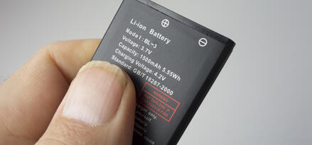 The image shows a close-up of a person's hand holding a lithium-ion (Li-ion) battery. The battery is labeled with technical specifications, including its model (BL-3), voltage (3.7V), capacity (1500mAh, 5.55Wh), and charging voltage (4.2V). There is a warning printed in red stating, "May explode if disposed in fire." The battery also displays positive and negative terminal symbols at the top. The background is a plain, light gray color, keeping the focus on the battery and hand.