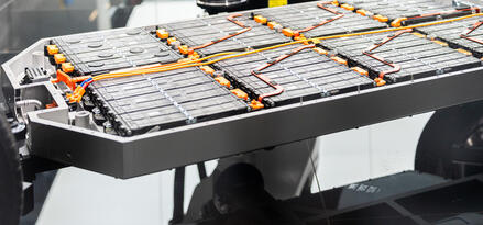 The image shows a close-up of an electric vehicle battery pack integrated into the chassis. The battery cells are organized in modules, with various orange high-voltage cables and connectors visible across the surface. A robotic arm is positioned above the battery pack, likely used for assembly or inspection purposes. In the background, you can see blurred automotive components, such as wheels and parts of vehicles, indicating a manufacturing or testing environment. The setup highlights advanced technology and precision engineering in electric vehicle production.