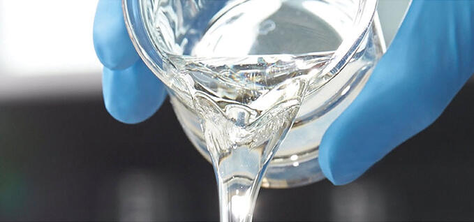 A close-up shows a clear, viscous liquid being poured from a glass beaker. The beaker is held by gloved hands wearing bright blue nitrile gloves, suggesting a lab or cleanroom setting. The liquid forms a smooth stream with glossy reflections. The background is softly blurred, keeping focus on the pouring action and laboratory glassware.
