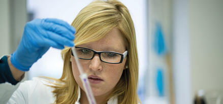 A blonde woman wearing glasses and a white lab coat stands in a laboratory. She is wearing light blue gloves and holding a syringe-like instrument, injecting liquid into a small vial.