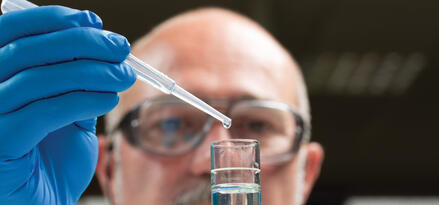 The image shows a person wearing blue gloves and holding a small transparent vial. The vial is likely used in a laboratory setting for scientific purposes, such as storing samples or conducting experiments. The focus on the gloves and vial suggests a controlled and sterile environment. The background appears to be a typical lab or office setting with ceiling lights visible.
