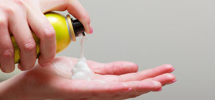 The image shows a person dispensing shaving foam from a yellow aerosol can onto their palm. The foam is white and fluffy, forming a mound as it is released. The can has some text visible on it, indicating it may contain product information or branding. The person’s hand is positioned to catch the foam, suggesting preparation for shaving. The background is plain and unobtrusive, focusing attention on the hands and the foam.